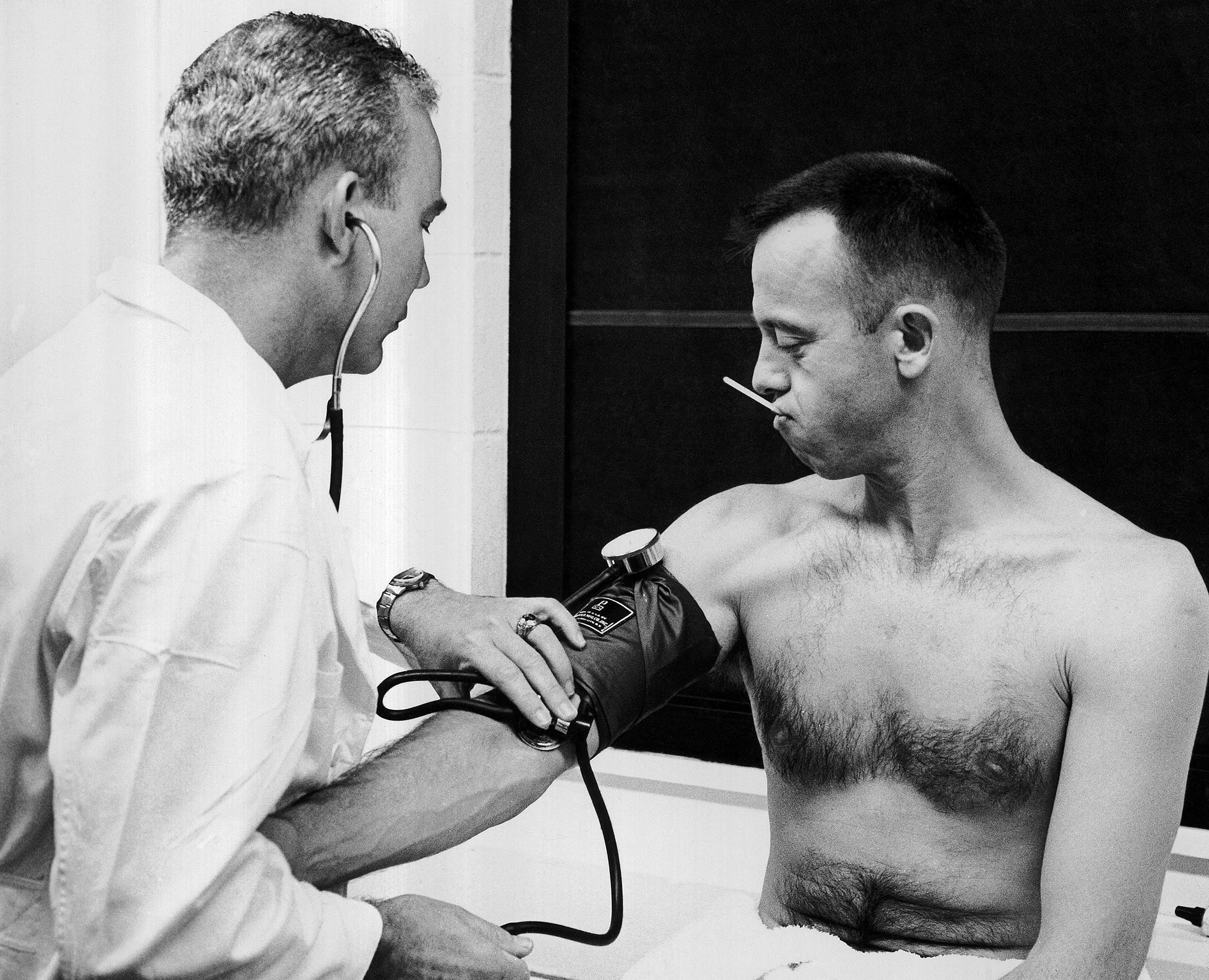 A thermometer in his mouth, Alan B. Shepard, Jr., watches as the astronaut's personal physician, Lt. Col. William K. Douglas, checks his blood pressure in Cape Canaveral, May 5, 1961. Shepard and his backup pilot, John H. Glenn, Jr., were given final physical examinations this morning at Cape Canaveral before Shepard took off on America's first manned space flight. (AP Photo)